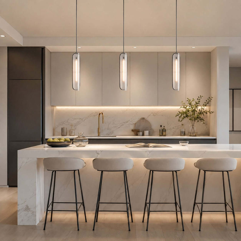 Row of three Clérachic capsule pendants lit above a marble kitchen island with four slim bar stools and under-cabinet lighting.