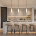 Row of three Clérachic capsule pendants lit above a marble kitchen island with four slim bar stools and under-cabinet lighting.