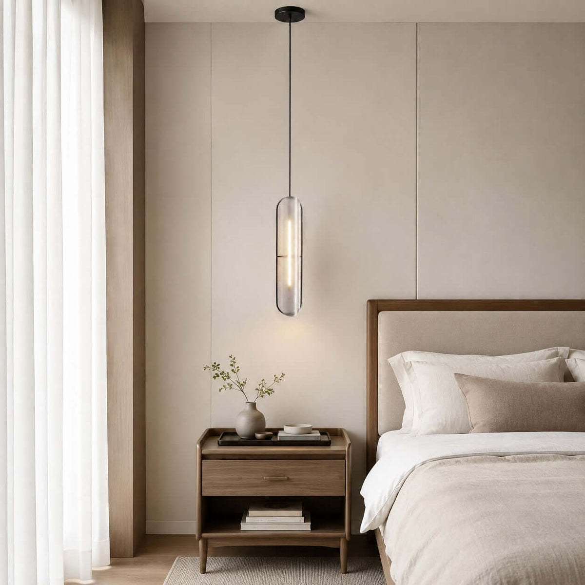 This Clérachic capsule pendant hangs by a bed with a wooden headboard, beside a bedside chest, a plant and sheer curtains.