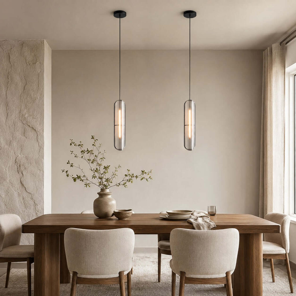 Pair of Clérachic capsule pendants on black cords over a wood dining table with boucle chairs, against a pale stone wall.