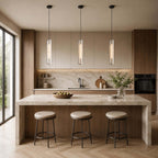 Three Clérachic capsule pendants lit over a travertine kitchen island with round leather stools and a herringbone floor.