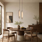 Two Clérachic capsule pendants at different heights above a round dining table with walnut chairs and a framed abstract artwork.