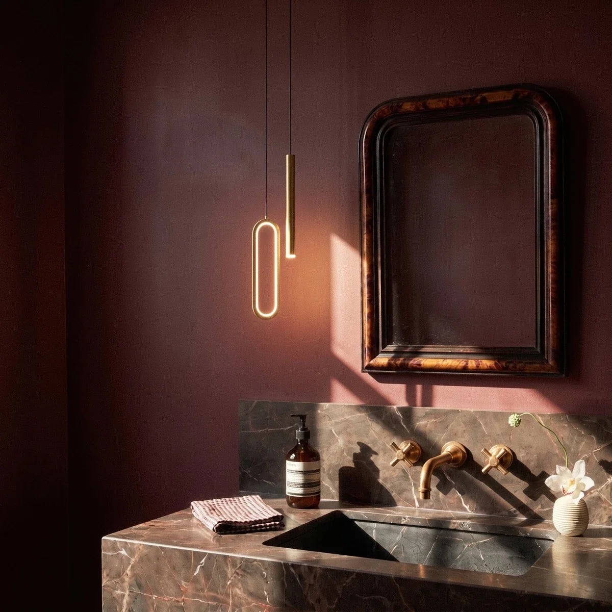 In a dark powder room, the brass pendant hangs beside an antique mirror above a marble vanity, Clérachic lifestyle shot.