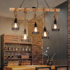 This Clérachic pendant light hangs over a wooden table in front of a brick wall, with five lit filament bulbs in black cages.