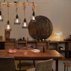 A Clérachic pendant light with caged bulbs on hemp rope, above a long dining table with a round wooden panel on the wall.