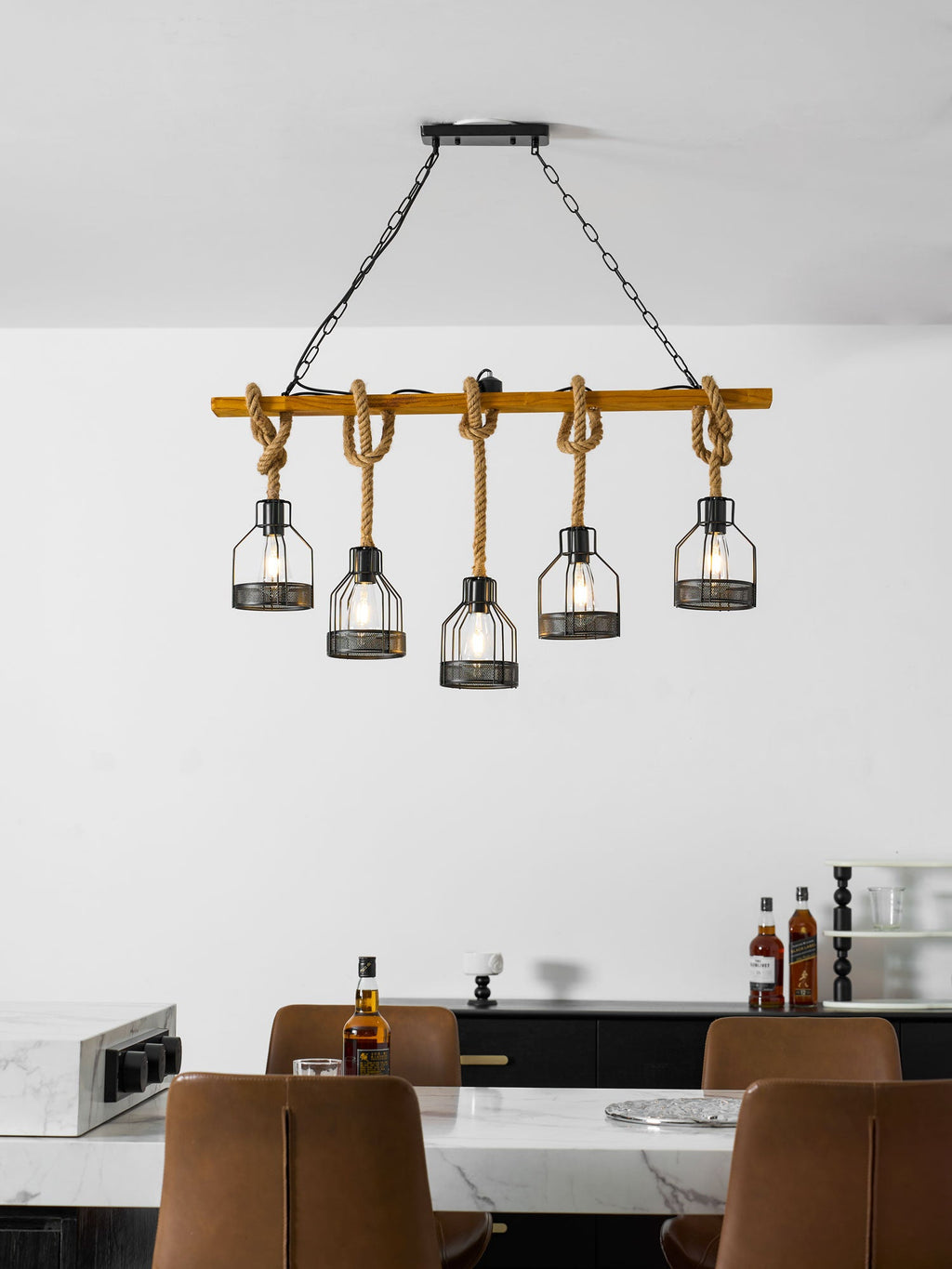 A Clérachic pendant light with five lit caged bulbs above a marble-topped counter with tan leather chairs and a bottle of whisky.