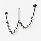 A Clérachic linear pendant light in black, with small glowing cylinders along a looping run hung from two ceiling plates, on white.