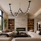 A Clérachic black linear pendant spans a living room in front of a stone fireplace, flanked by shelves and two cream sofas.