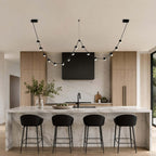 A Clérachic linear pendant with small round lit beads sweeps over a marble kitchen island with four black bar stools.
