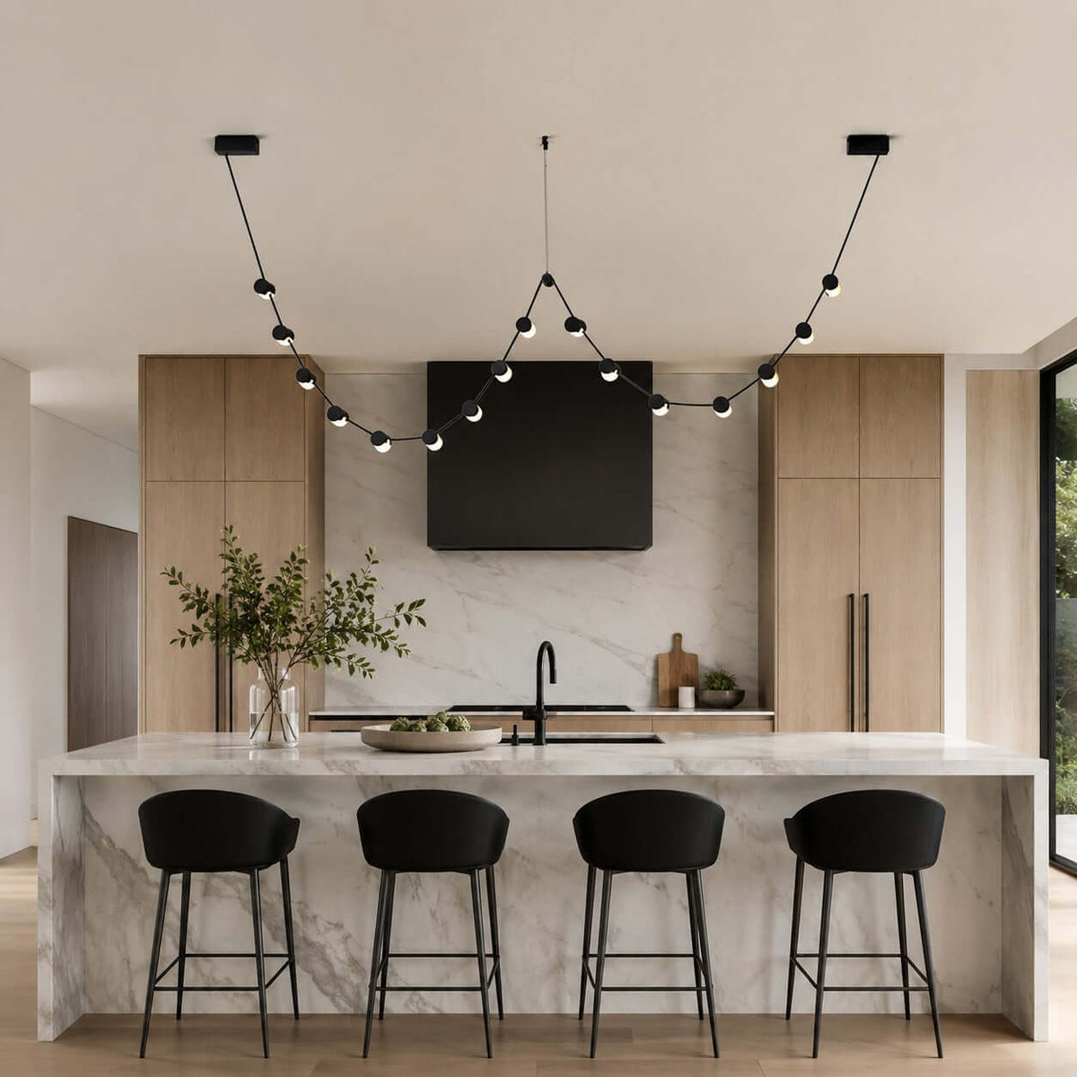A Clérachic linear pendant with small round lit beads sweeps over a marble kitchen island with four black bar stools.