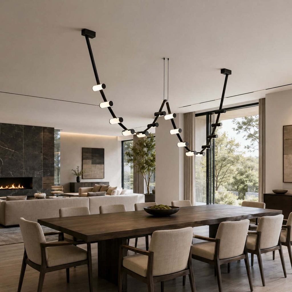 A Clérachic linear pendant arcs over a dark dining table with arm chairs, in a room with a fireplace and a wall of glass.