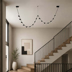 Black Clérachic beaded pendant light looping across the ceiling of a stairwell, beside a black stair rail and a pale wall.