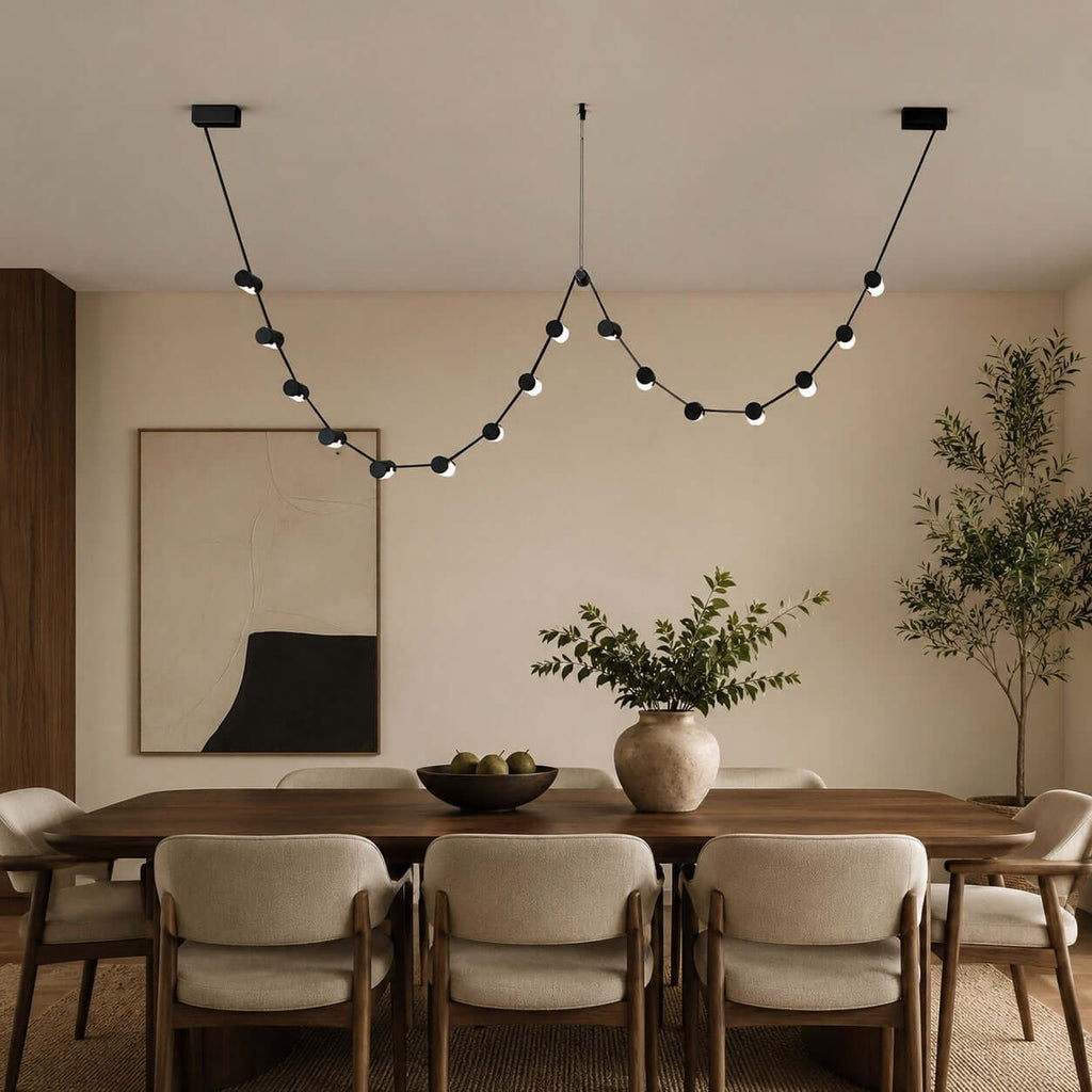 This Clérachic pendant light, a string of small black beads in two loops, hangs above a dining table with upholstered chairs.