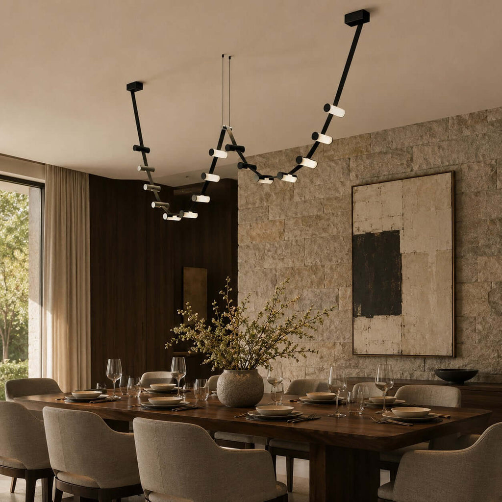 A Clérachic linear pendant lit above a set dining table, with a stone feature wall, cream chairs and a vase of branches.