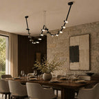 A Clérachic linear pendant lit above a set dining table, with a stone feature wall, cream chairs and a vase of branches.