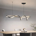Clérachic long ribbon pendant with arching black loops lit warm above a pale dining table, chairs and a small clock on books.