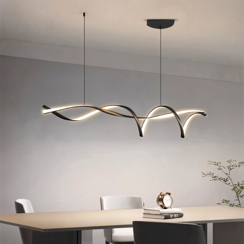 Clérachic long ribbon pendant with arching black loops lit warm above a pale dining table, chairs and a small clock on books.