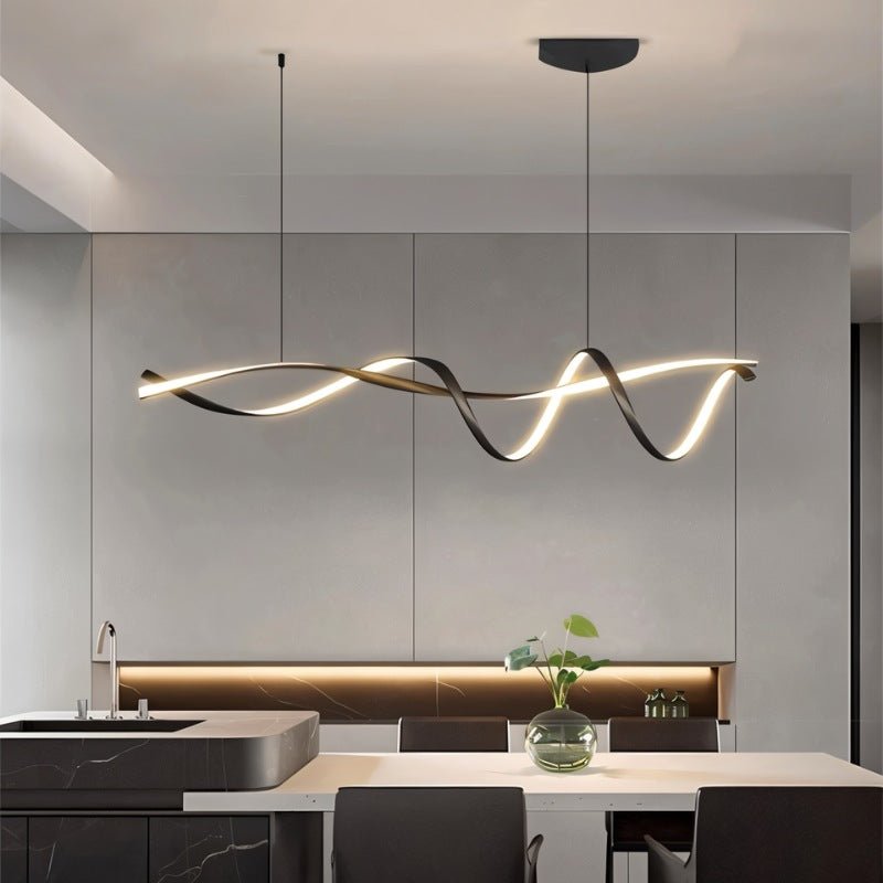 Clérachic glowing ribbon pendant above a kitchen island and dining table, with a glass vase, dark chairs and a marble-veined worktop.