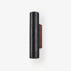 A black travertine cylinder wall light on a walnut backplate, switched off against a plain white backdrop, from Clérachic.
