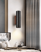 This Clérachic wall light casts a soft pool of warm light downward beside grey curtains and a bed with a throw.