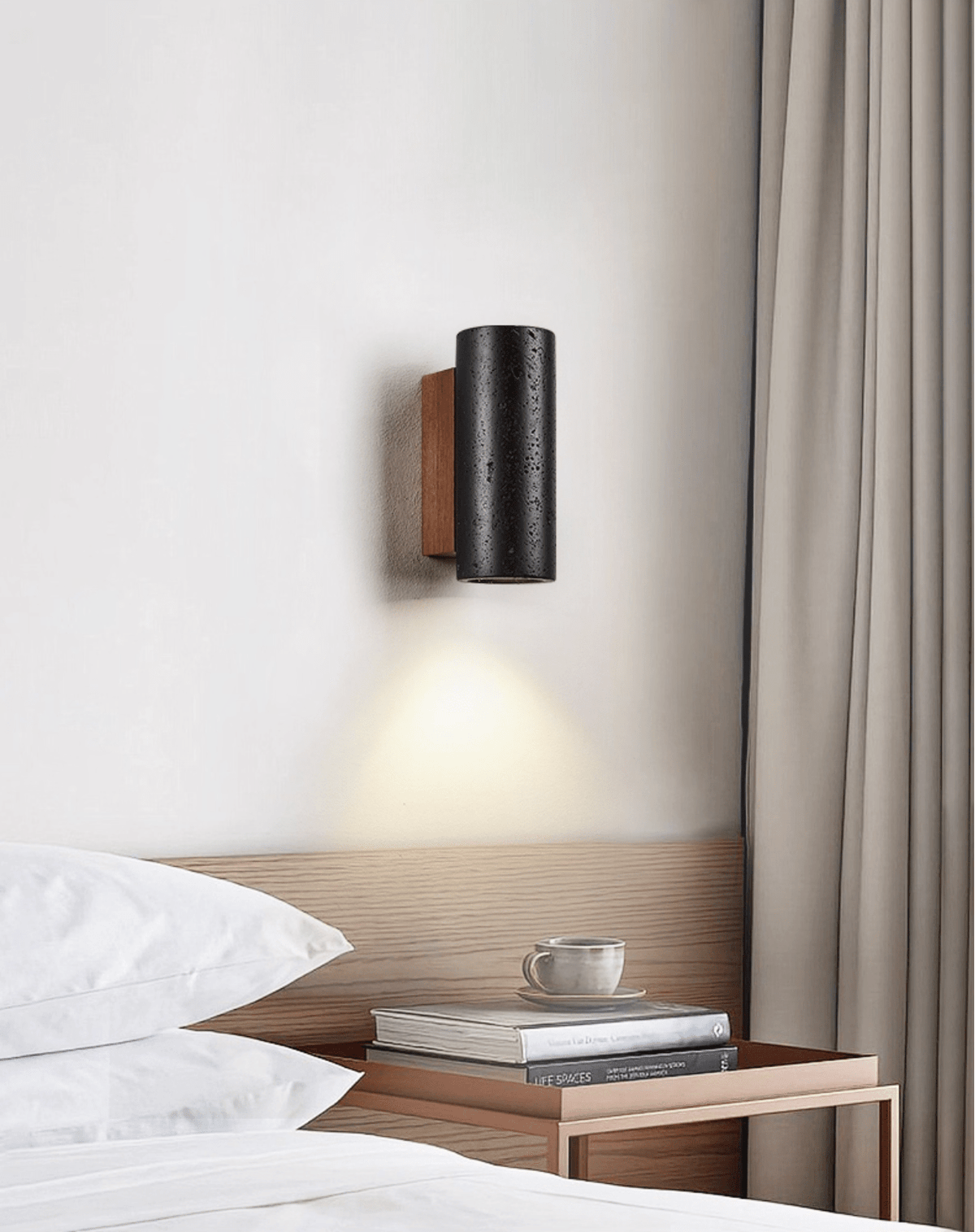 A short black Clérachic cylinder wall light with a walnut plate shines down above a bedside table and white pillows.