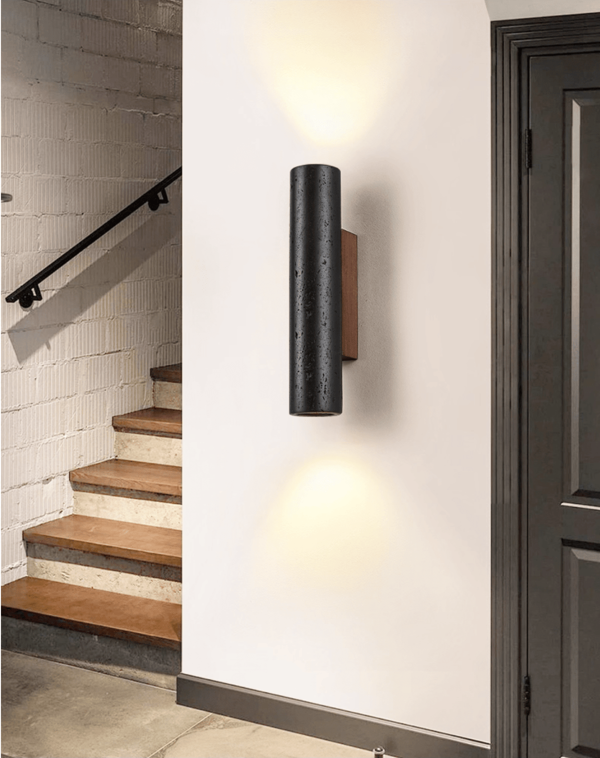 Light spills up and down from a Clérachic black stone wall light beside a dark doorway and a timber staircase.