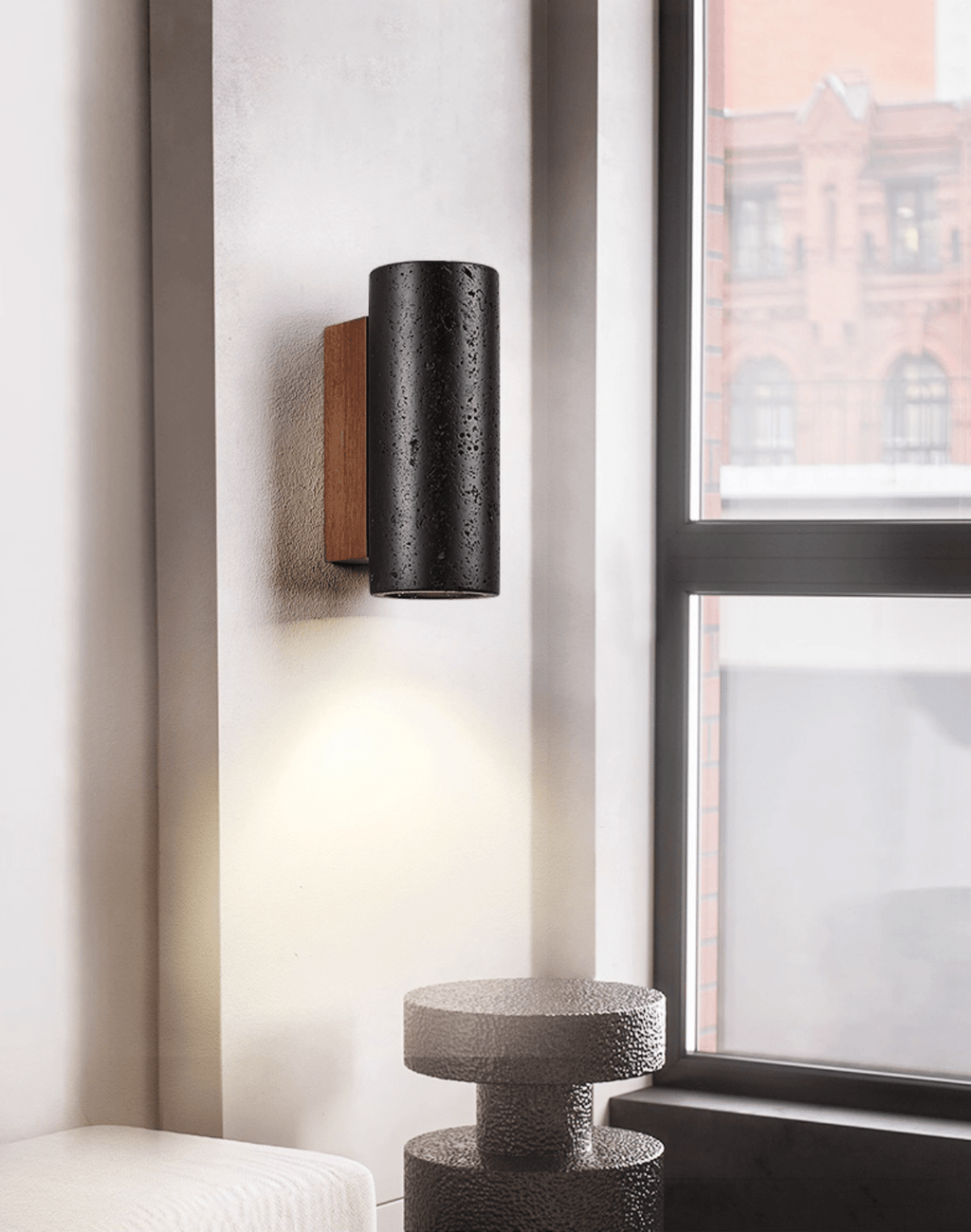 A black travertine Clérachic wall light glows downward by a tall window, above a rough stone side table.