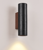 Close view of a Clérachic black travertine cylinder with pitted texture, a warm glow below it and a walnut plate behind.