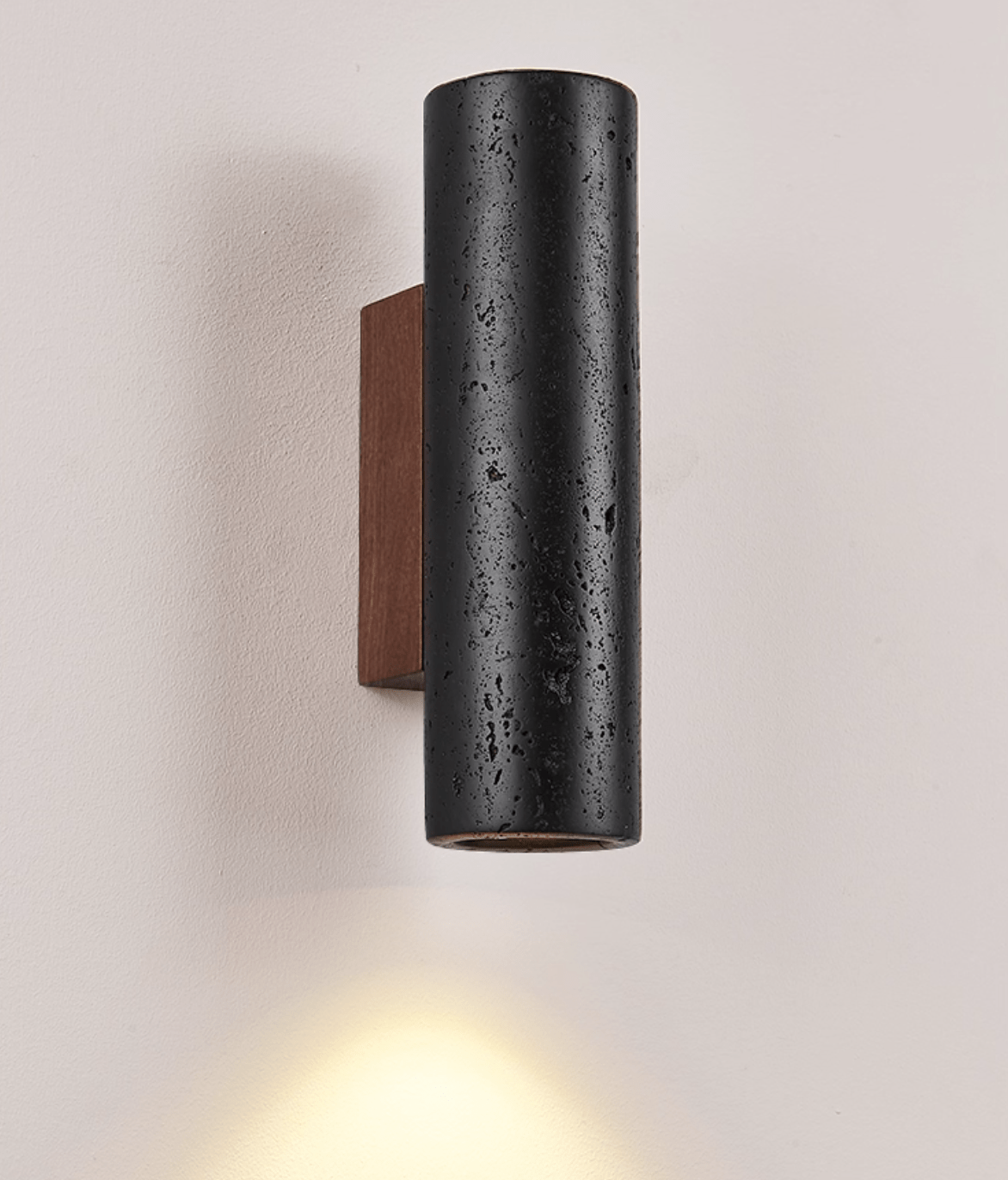 Close view of a Clérachic black travertine cylinder with pitted texture, a warm glow below it and a walnut plate behind.