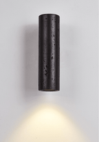 A Clérachic black stone cylinder light mounted on a pale wall, throwing a warm fan of light down to the floor.