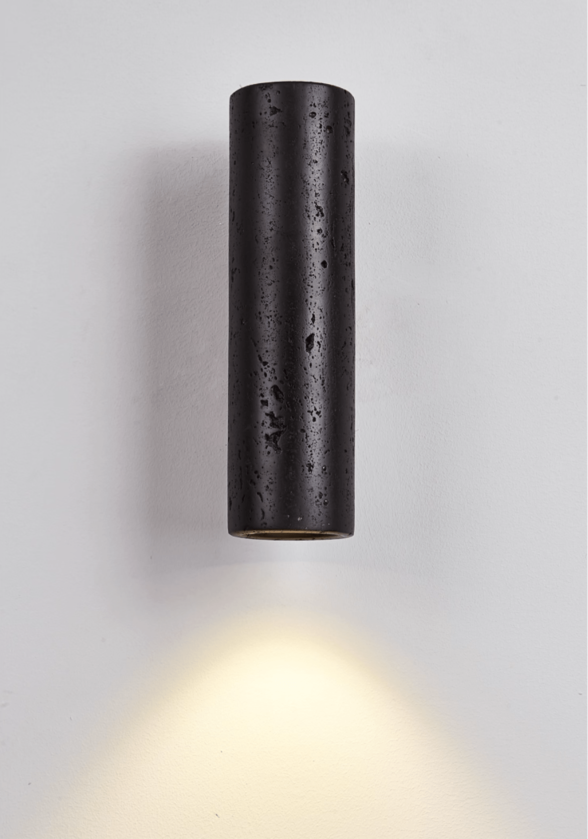 A Clérachic black stone cylinder light mounted on a pale wall, throwing a warm fan of light down to the floor.