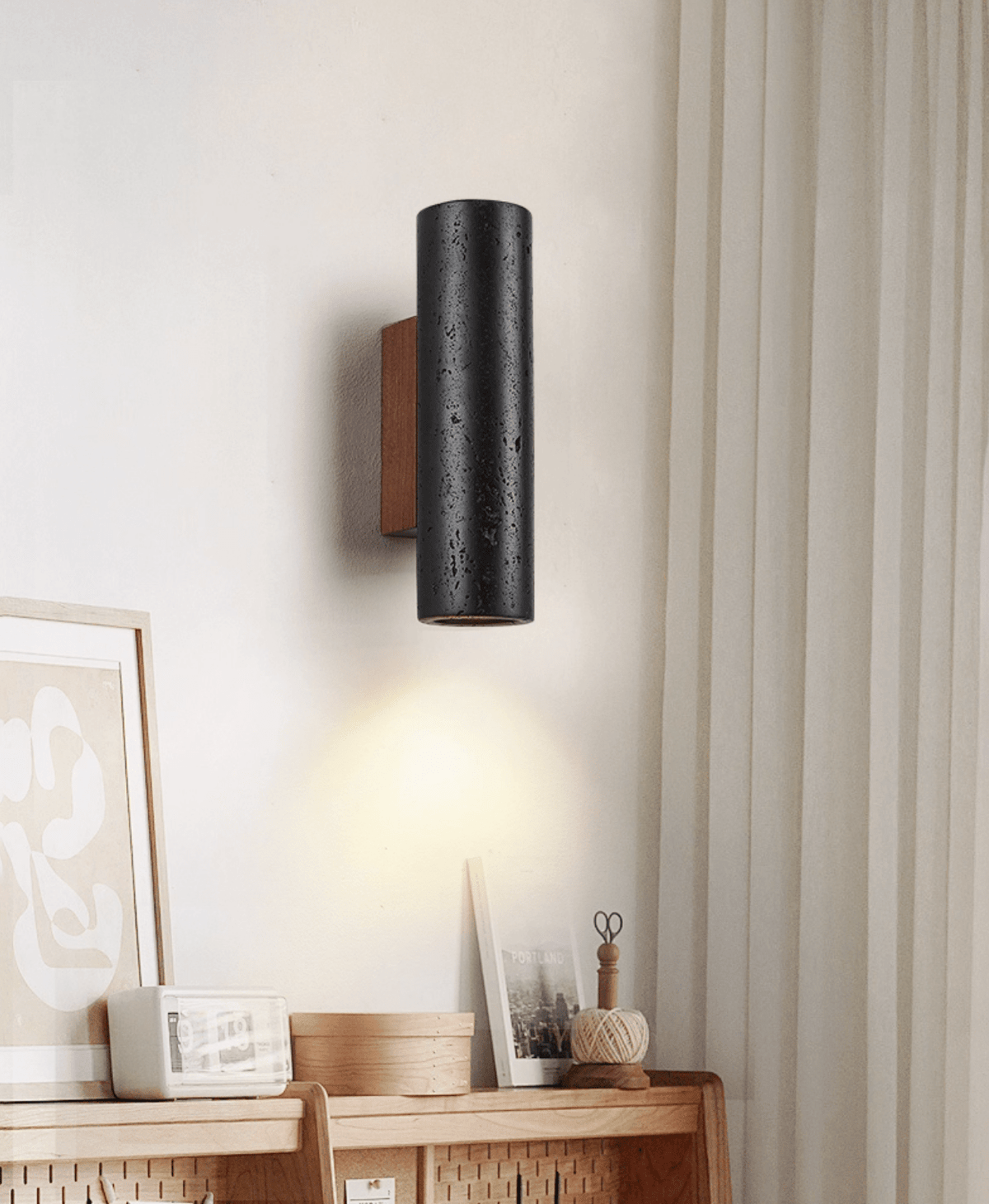A Clérachic cylinder wall light with a walnut plate lights a sideboard with framed art, a small clock and a basket.