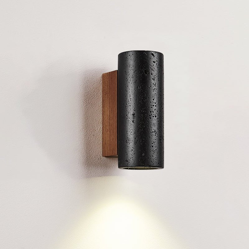 Close view of a short Clérachic black travertine cylinder on its walnut plate, light pooling down the wall below.