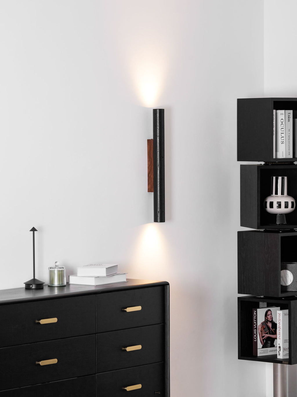 A tall Clérachic cylinder wall light lights the wall above a black sideboard and a stack of dark cube shelves.