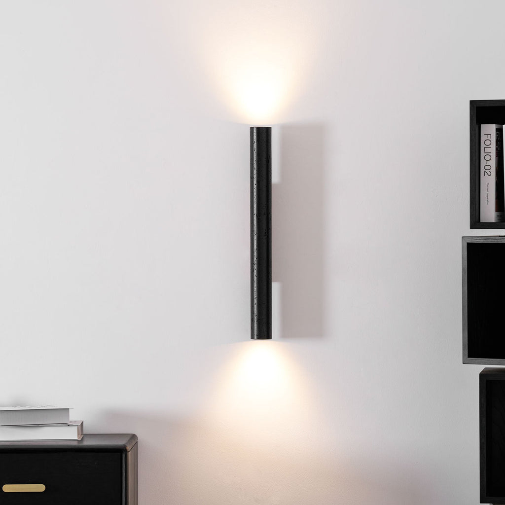 A slim Clérachic cylinder wall light glowing up and down on a white wall, with a black sideboard and shelves nearby.