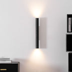 A slim Clérachic cylinder wall light glowing up and down on a white wall, with a black sideboard and shelves nearby.
