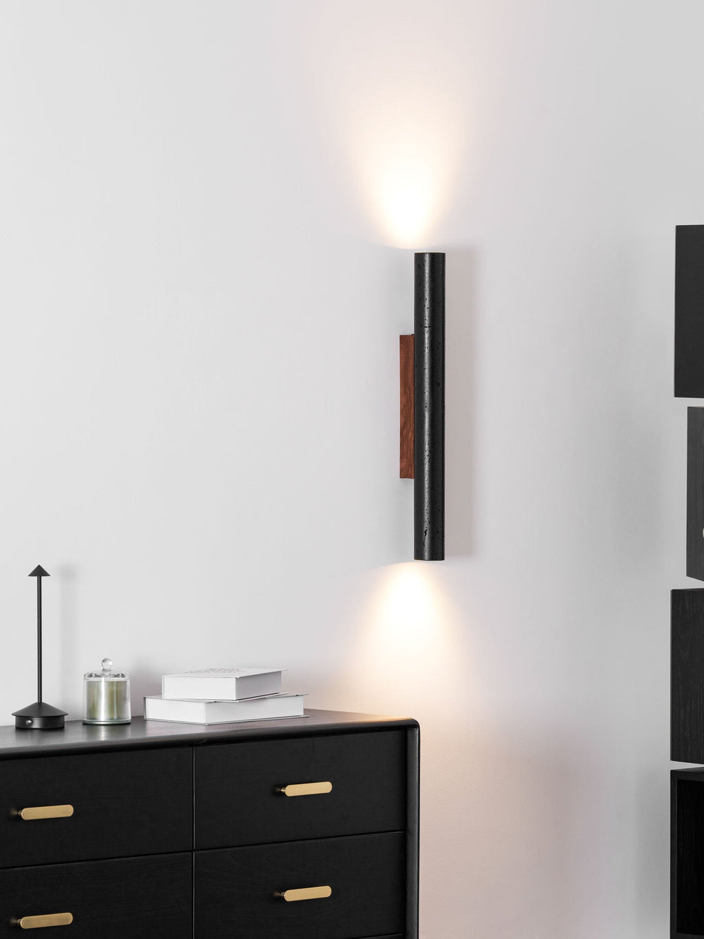 A tall Clérachic wall light with a walnut backplate casts light above and below, with a black chest and a small table lamp.