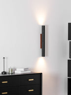 A tall Clérachic wall light with a walnut backplate casts light above and below, with a black chest and a small table lamp.