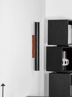 A tall Clérachic black cylinder with a walnut plate stands out on a white wall beside a stack of dark open shelves.