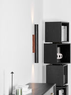 A tall Clérachic cylinder wall light with a faint glow, beside black cube shelves holding books and a striped vase.