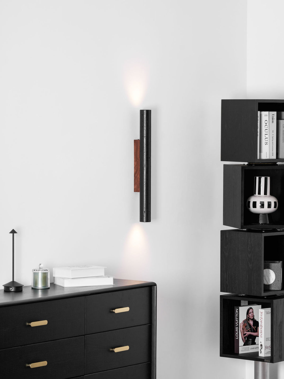A slender Clérachic wall light glows softly up and down above a black chest of drawers and a small cordless lamp.