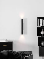 A tall Clérachic cylinder wall light glowing both ways above a black leather armchair and a dark sideboard.