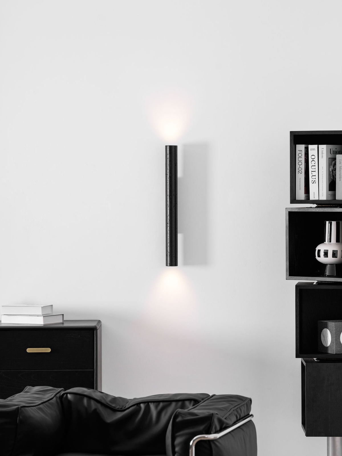 A tall Clérachic cylinder wall light glowing both ways above a black leather armchair and a dark sideboard.