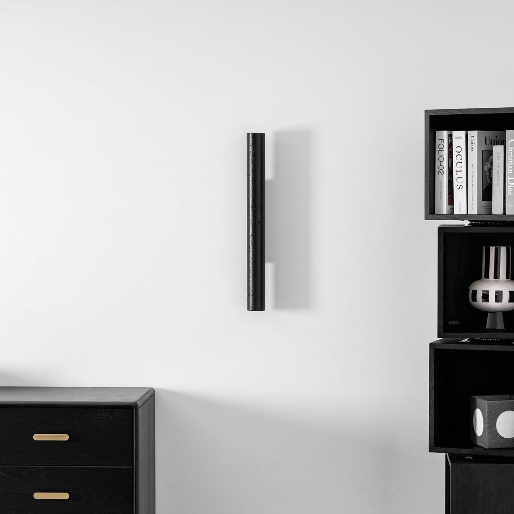A tall Clérachic black cylinder, unlit, on a white wall between a black chest of drawers and open cube shelves.