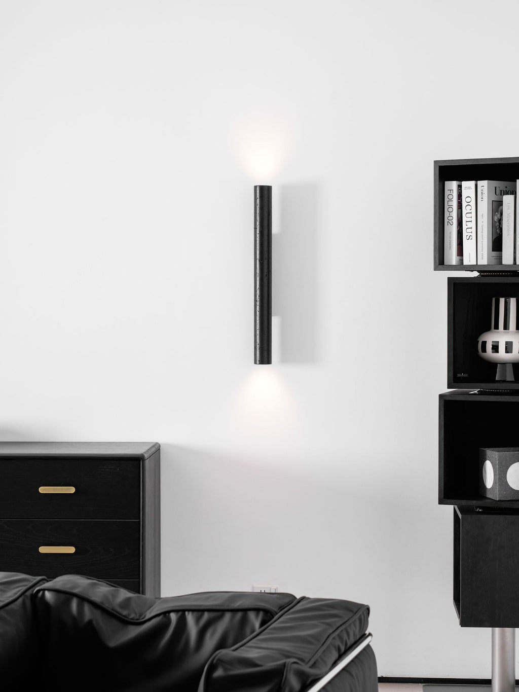 A tall Clérachic cylinder wall light with a soft up and down glow, above a black armchair and a chest with brass pulls.