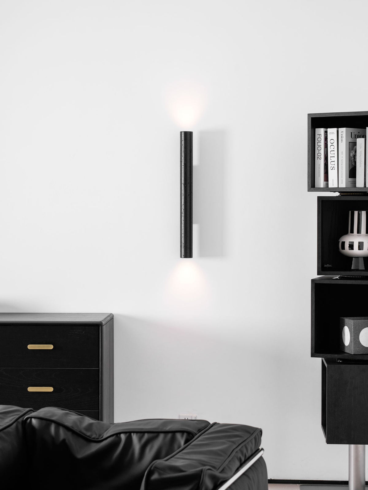A tall Clérachic cylinder wall light with a soft up and down glow, above a black armchair and a chest with brass pulls.