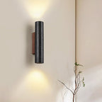 A Clérachic cylinder wall light glows up and down on a cream wall beside a slender branch in the corner.