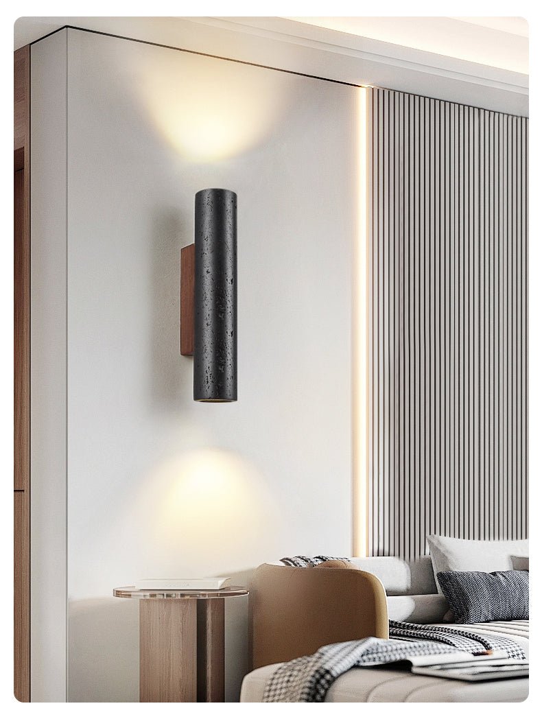 A Clérachic black cylinder wall light glows above a bedroom with a camel chair, a ribbed wall panel and cushions.