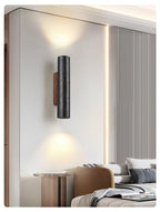 A Clérachic black cylinder wall light glows above a bedroom with a camel chair, a ribbed wall panel and cushions.