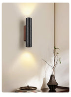 A Clérachic cylinder wall light lights a cream wall above a dark vase, a branch and a stack of books on a console.